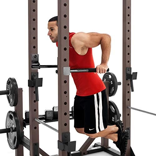 A muscular man in a red tank top performing weighted dips on the inside dip handles of the Steelbody Monster Cage, with loaded barbell plates stored on the side horns